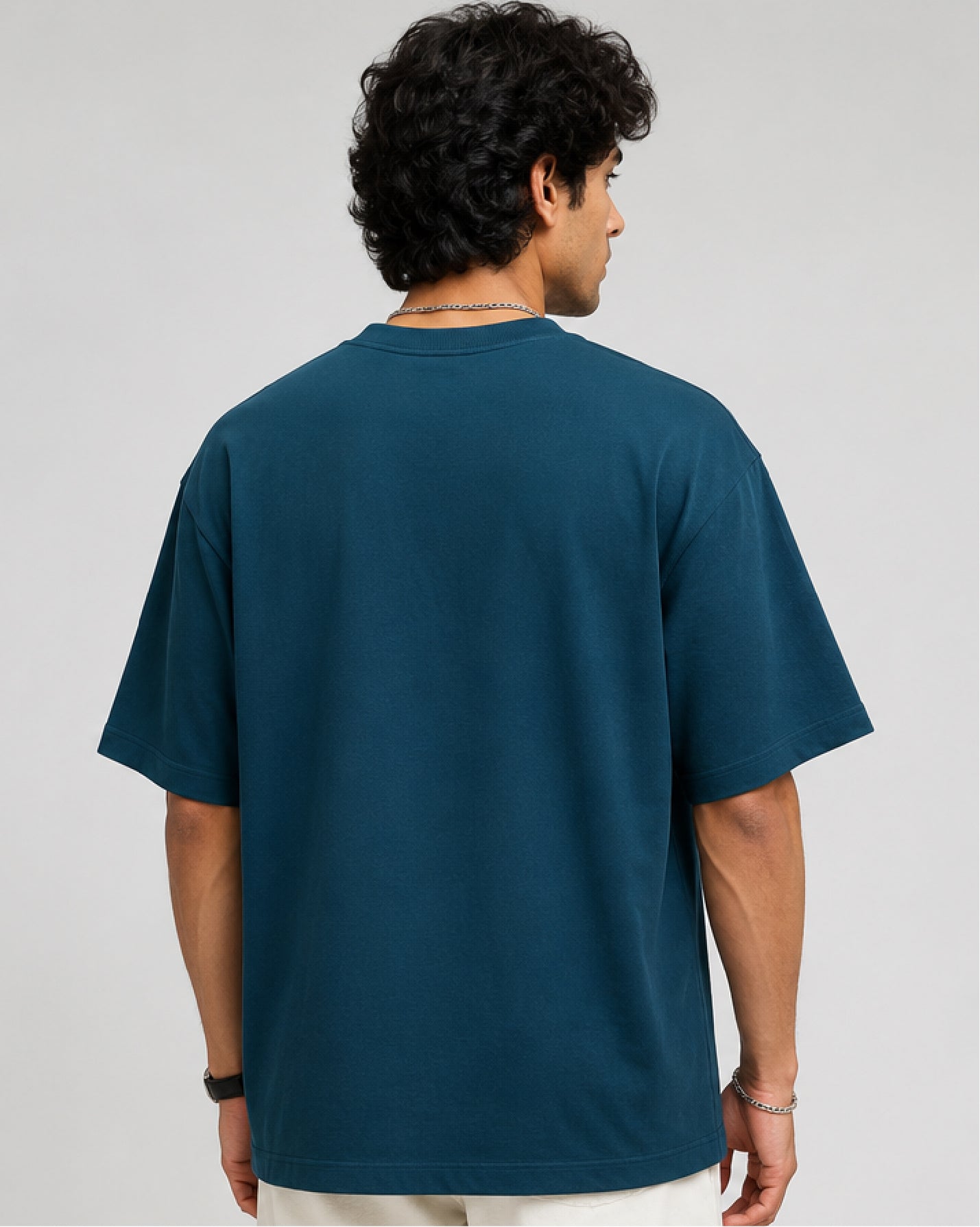 A man viewed from the back wearing an oversized dark teal blue t-shirt and light-colored pants, set against a plain light gray studio background.