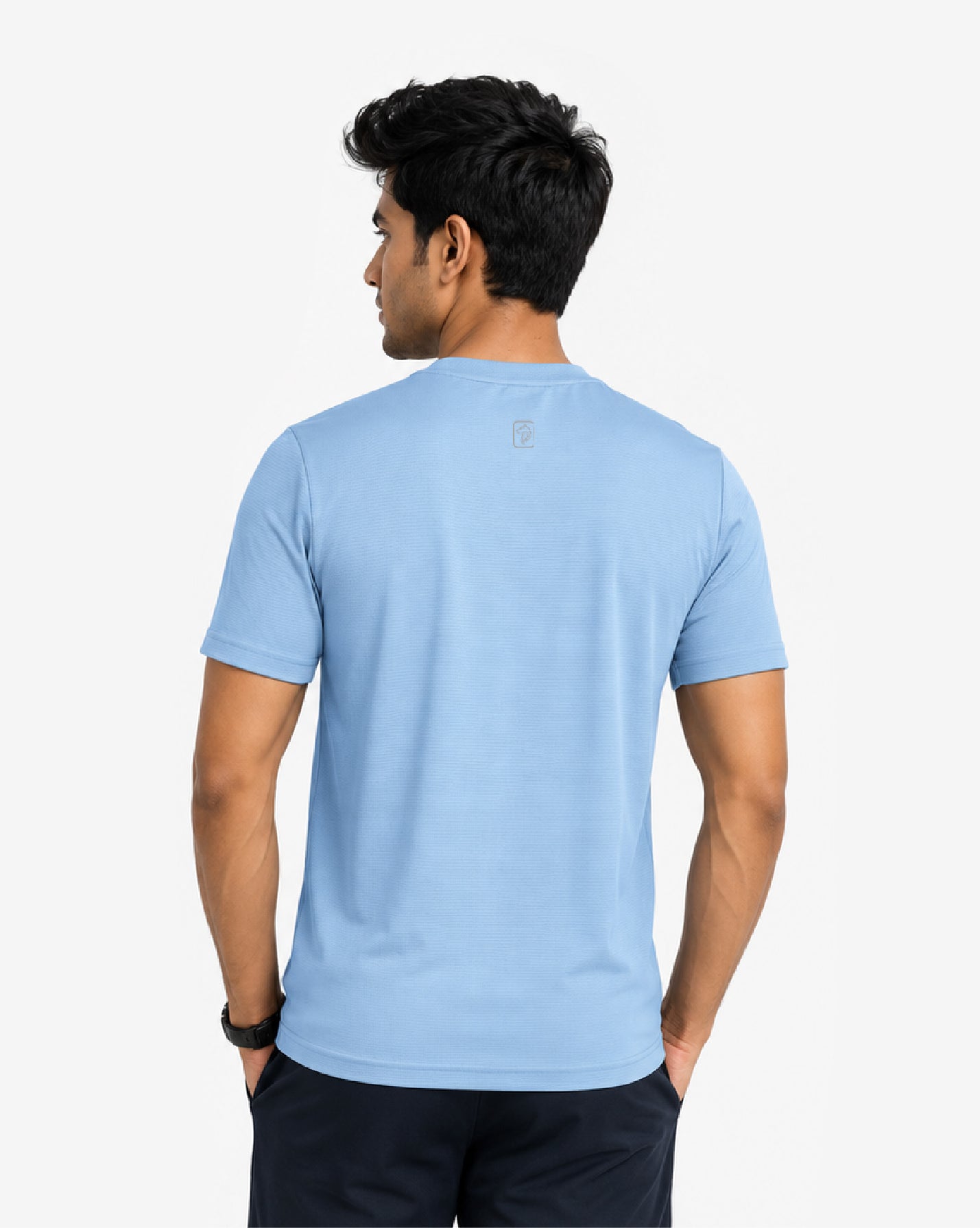 A man viewed from the back wearing a light blue short-sleeve crewneck t-shirt and dark navy pants, set against a plain light gray studio background.